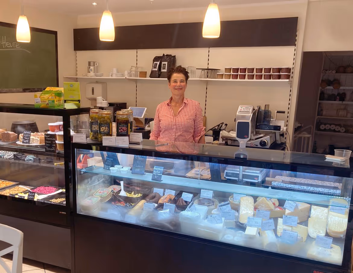 Lächelnde Frau in einem karierten Hemd steht hinter einer Vitrine mit Käse und anderen Produkten in einem kleinen Feinkostladen.